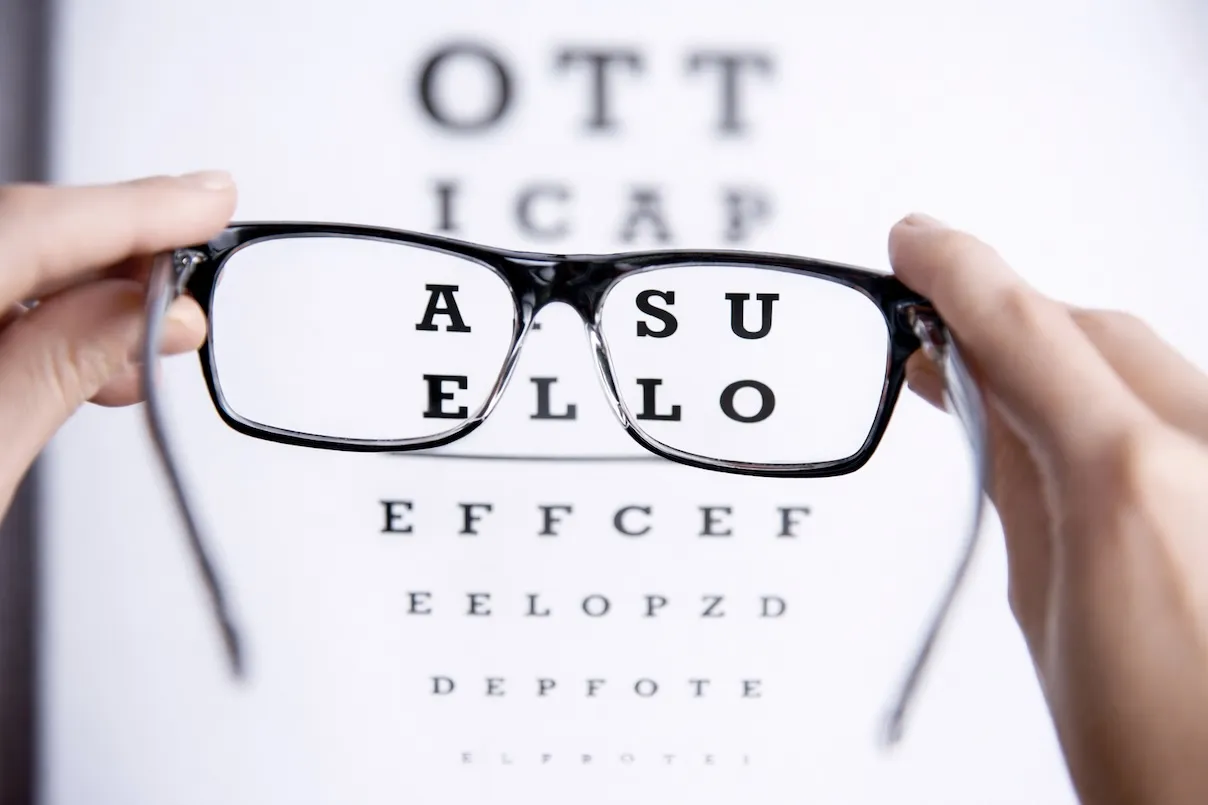 Occhiali neri davanti a un tabellone oculistico con le lettere 'OTT ICAP ASSU ELLO' nitide dentro le lenti e sfocate fuori.