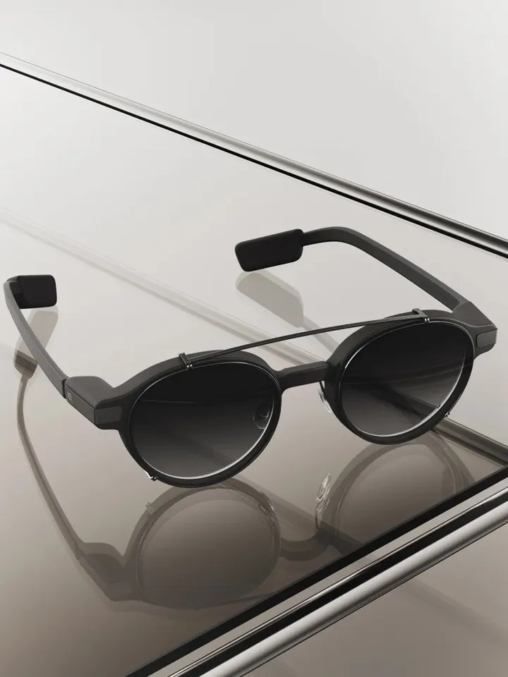 Occhiali smart dal design moderno con montatura nera e lenti scure appoggiati su una superficie riflettente.