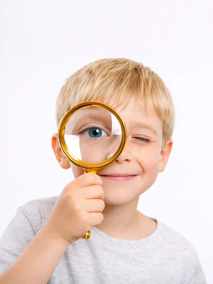 Bambino biondo sorride e guarda in camera tenendo una lente d’ingrandimento davanti all’occhio destro, che appare ingrandito.